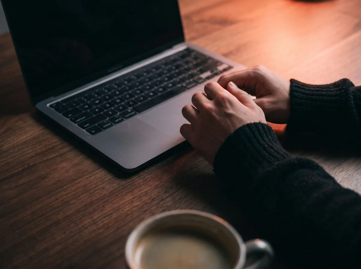 Des mains posées à côté du clavier d’un MacBook sur un bureau sombre.